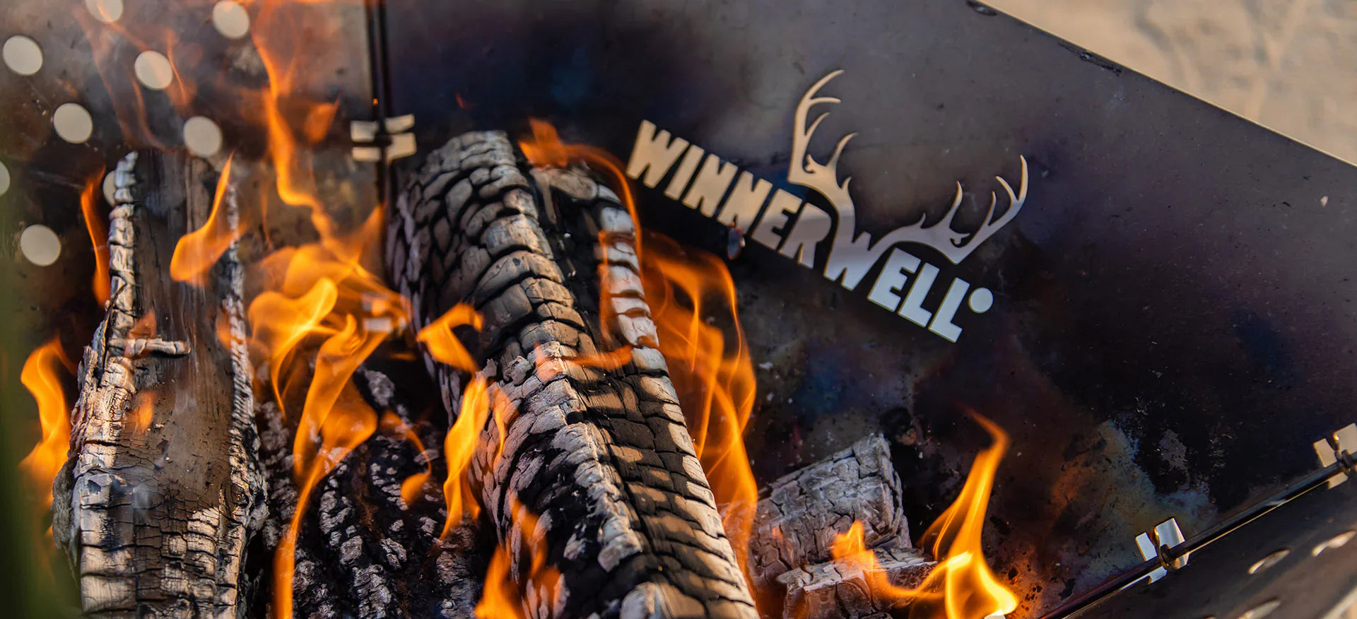Brandende houtblokken met oranje vlammen in een metalen vuurkorf met het uitgesneden logo “WINNERWELL” en een afbeelding van een hertengewei.