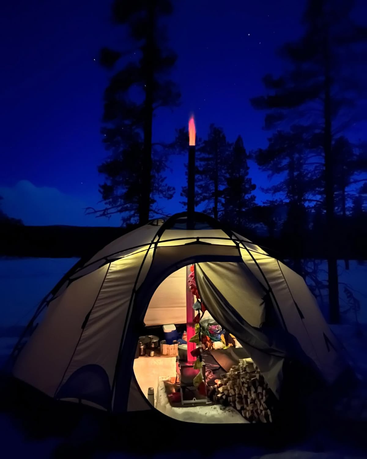 Zelt im Schnee bei Nacht mit dem Winnerwell Nomad View darin und einer Flamme aus dem Schornstein