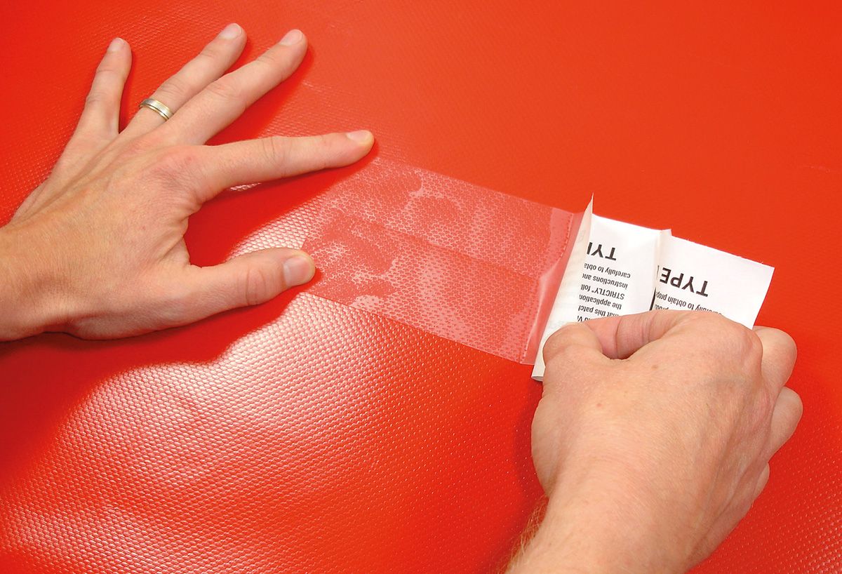Tear-Aid B Flicken wird mit zwei Händen auf rotes PVC-Gewebe geklebt