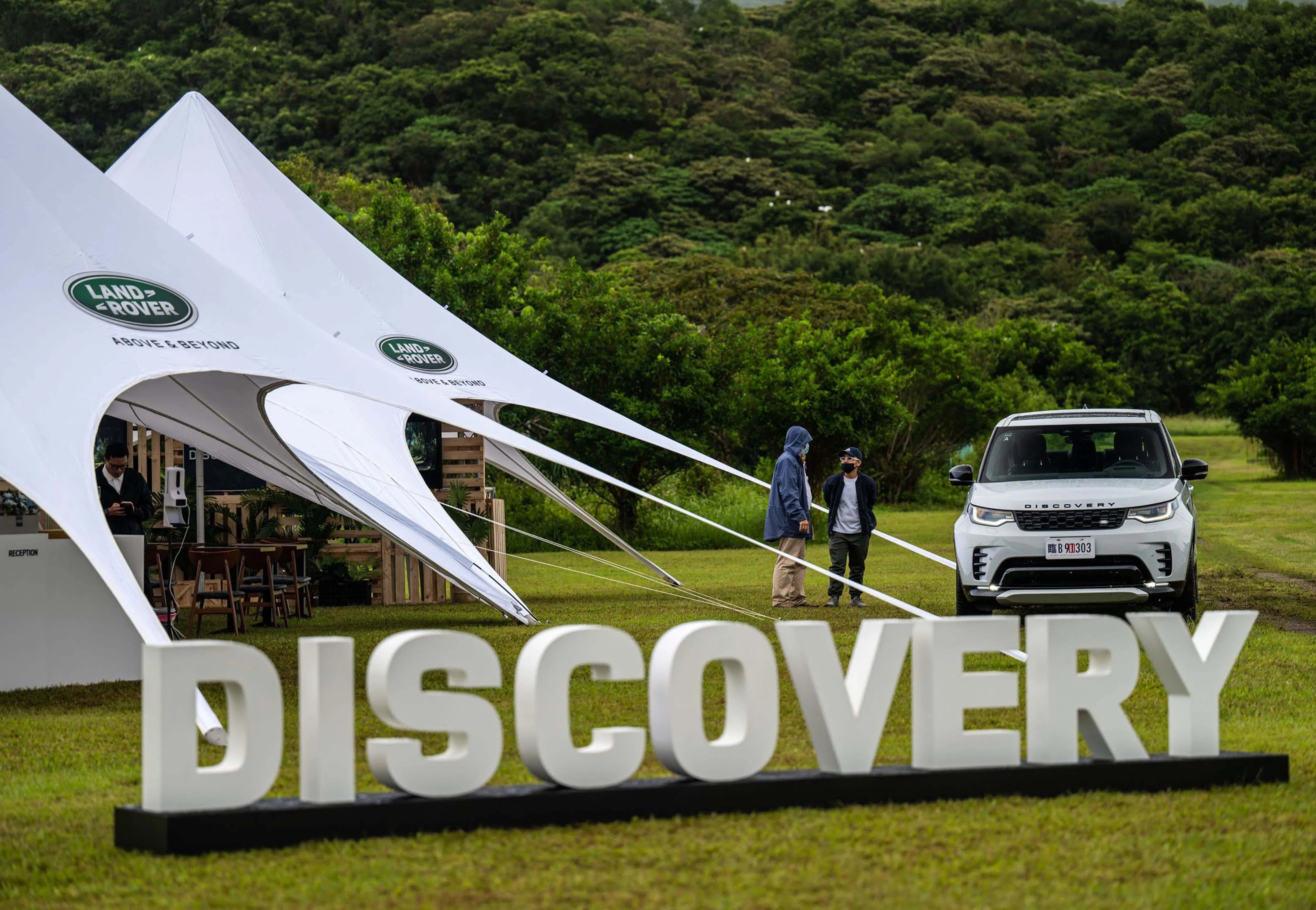 Evenement met grote witte tenten met het Land Rover-logo "Above & Beyond", opgezet op een grasveld omringd door bossen. 