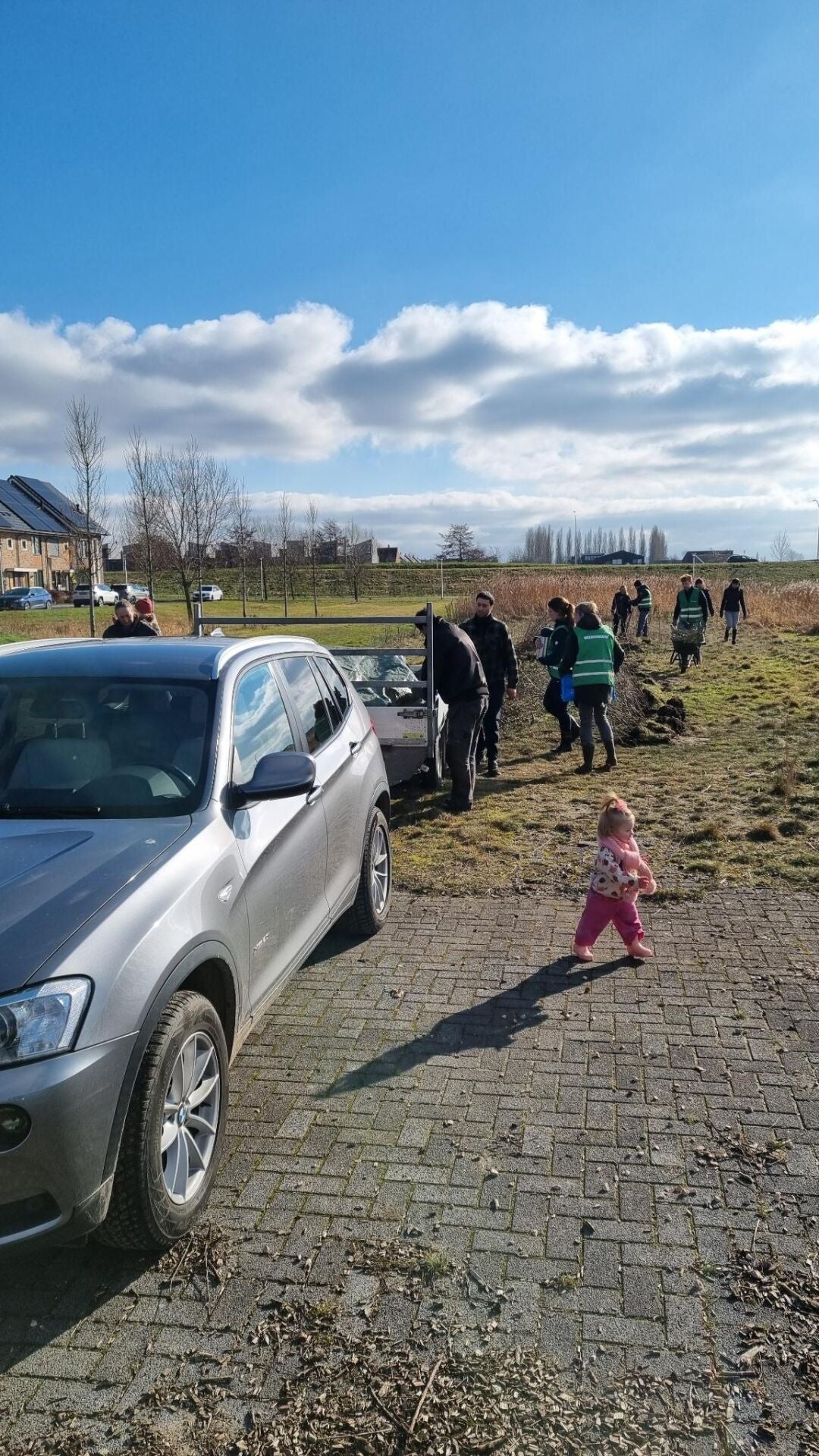 Een groep mensen staat en loopt in een open veld langs een rij pas geplante boompjes, naast een geparkeerde auto en een kleine aanhanger; op de voorgrond loopt een klein kind over een stenen pad, met huizen en een brede lucht met wolken op de achtergrond.
