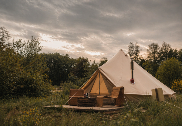 Beige Bell Tent 500 op een houten platform in een groene, bosrijke omgeving, ingericht als glampingplek met rotan stoelen en een klein tafeltje, onder een bewolkte lucht met warm avondlicht.