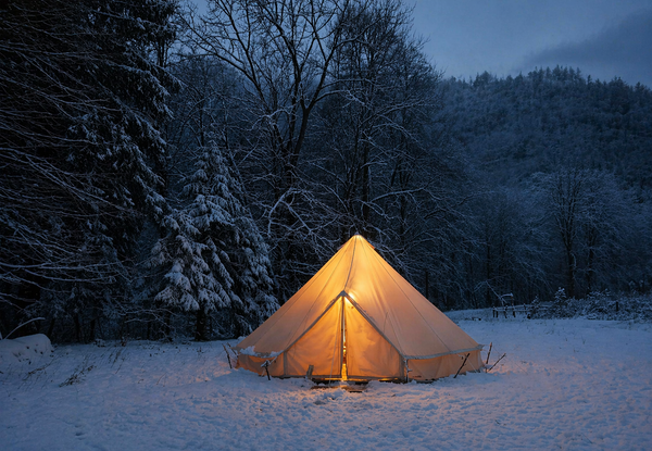 Beige Bell Tent 600 verlicht van binnen in een besneeuwd boslandschap tijdens de schemering, omringd door met sneeuw bedekte bomen en een blauwe, winterse sfeer.