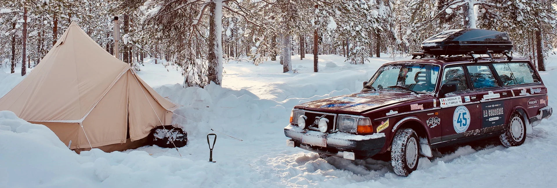Beige tent opgezet in een besneeuwd bos naast een donkerrode rallyauto met dakdrager, stickers en het nummer 45 op de deur; een schep staat rechtop in de sneeuw ertussenin.
