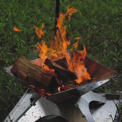 platte metalen firepit met brandende houtblokken en hoge vlammen, geplaatst op gras in een buitenomgeving