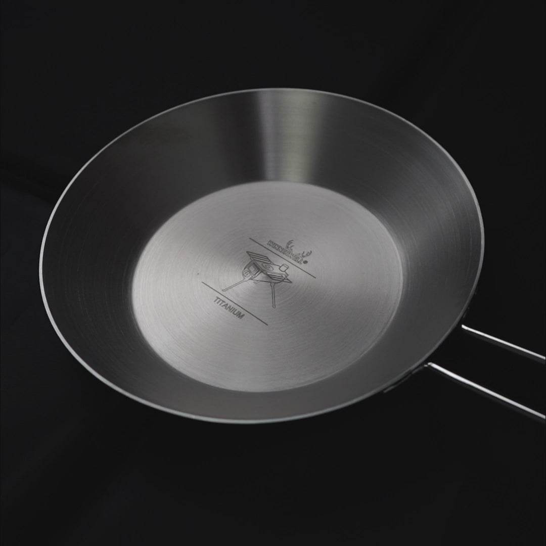 titanium koekenpan met opklapbare handgreep en gegraveerd logo in het midden, weergegeven tegen een donkere achtergrond