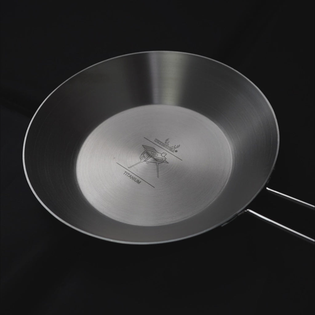titanium koekenpan met opklapbare handgreep en gegraveerd logo in het midden, weergegeven tegen een donkere achtergrond