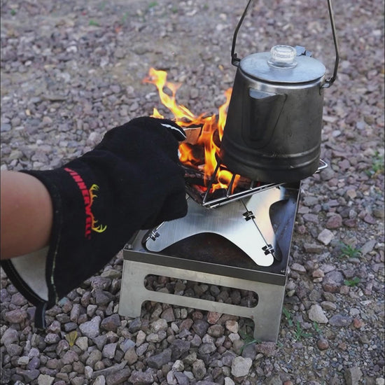 draagbare metalen firepit met open vuur terwijl een hand met hittebestendige handschoen hout toevoegt en een waterketel boven de vlammen hangt op een grindondergrond