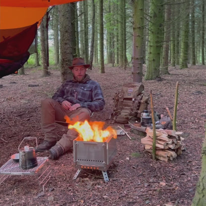 opvouwbare metalen campingkachel tijdens montage in het bos, terwijl een persoon de uitklapbare poten en zijpanelen installeert op de grond buiten