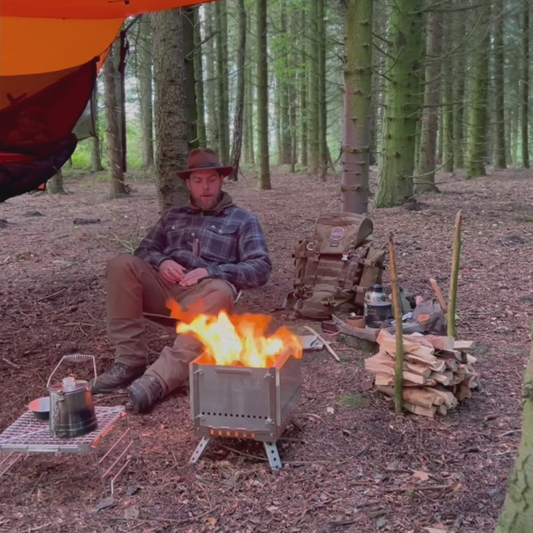 opvouwbare metalen campingkachel tijdens montage in het bos, terwijl een persoon de uitklapbare poten en zijpanelen installeert op de grond buiten