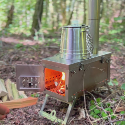 titanium camping houtkachel van Winnerwell met open vuurhaard en metalen ketel bovenop, gebruikt in een bosrijke kampeeromgeving