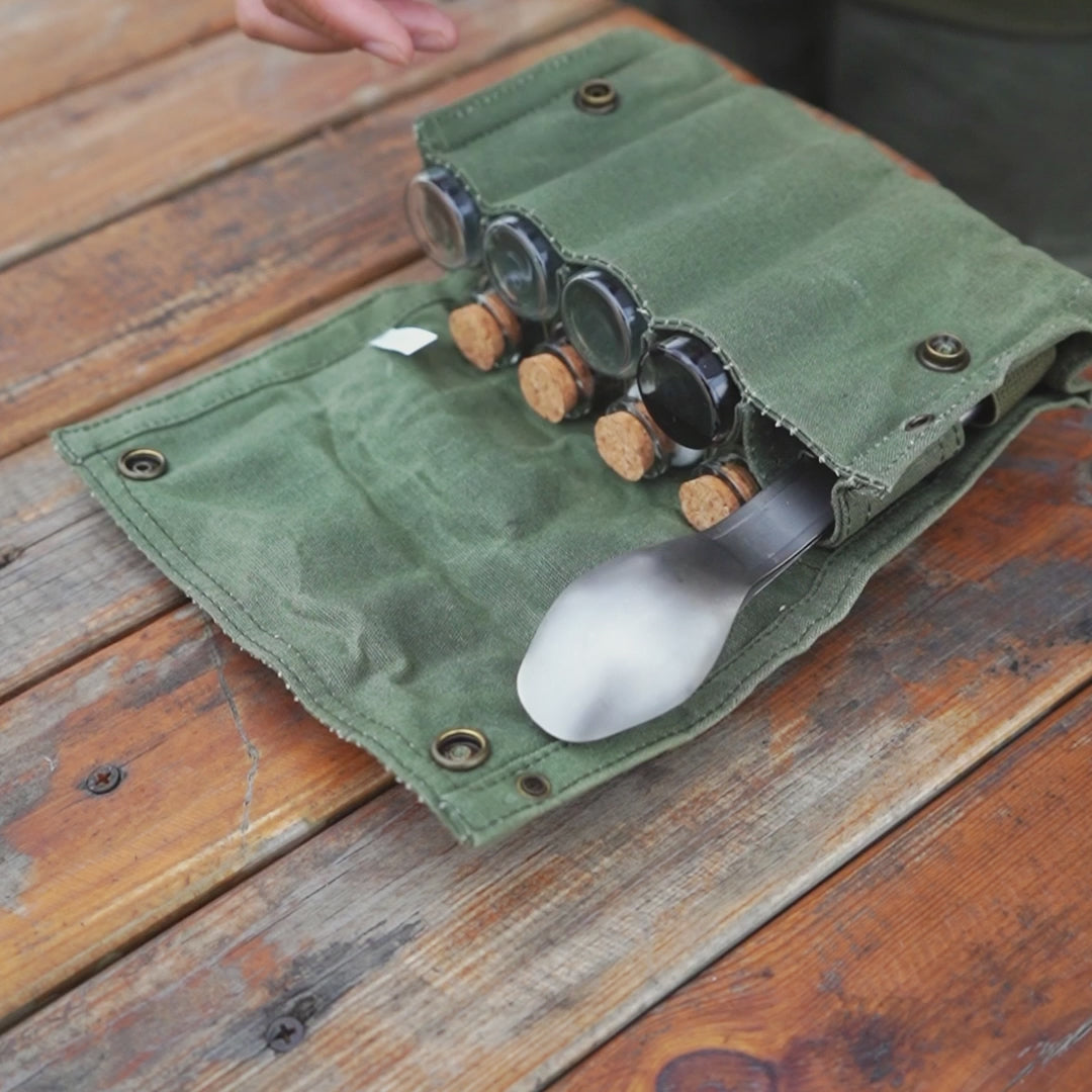 opgerolde groene hoes met titanium camping bestekset bestaande uit lepel, vork en mes, samen met kleine potjes met kurken doppen op een houten tafel