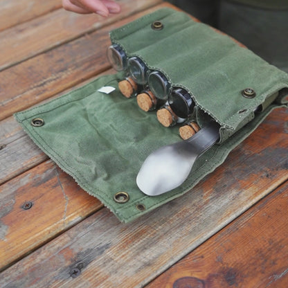 opgerolde groene hoes met titanium camping bestekset bestaande uit lepel, vork en mes, samen met kleine potjes met kurken doppen op een houten tafel