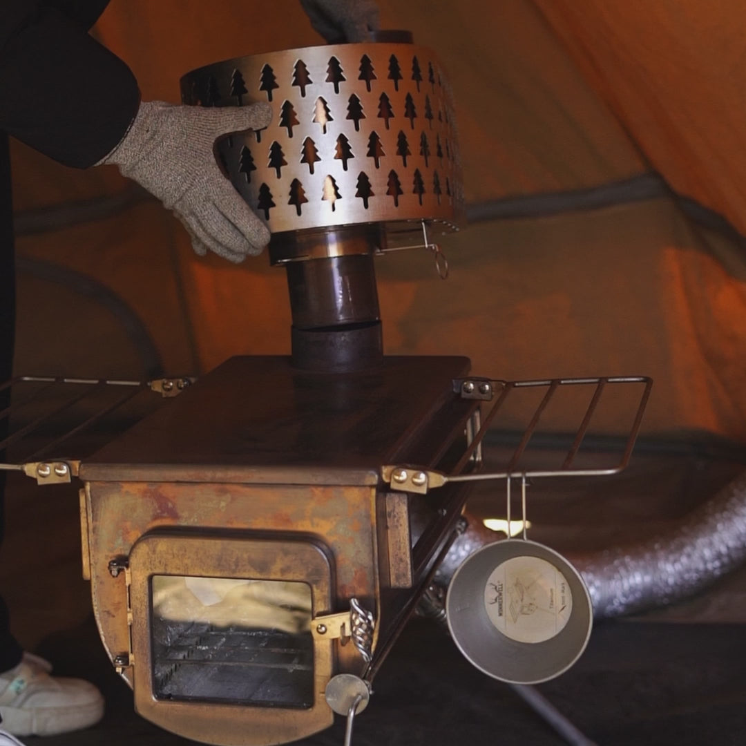 metalen vonkvanger met boompatroon die op een kachelpijp wordt geplaatst terwijl iemand met handschoenen deze vastzet, bovenop een houtkachel met zichtbaar raam en zijrekken