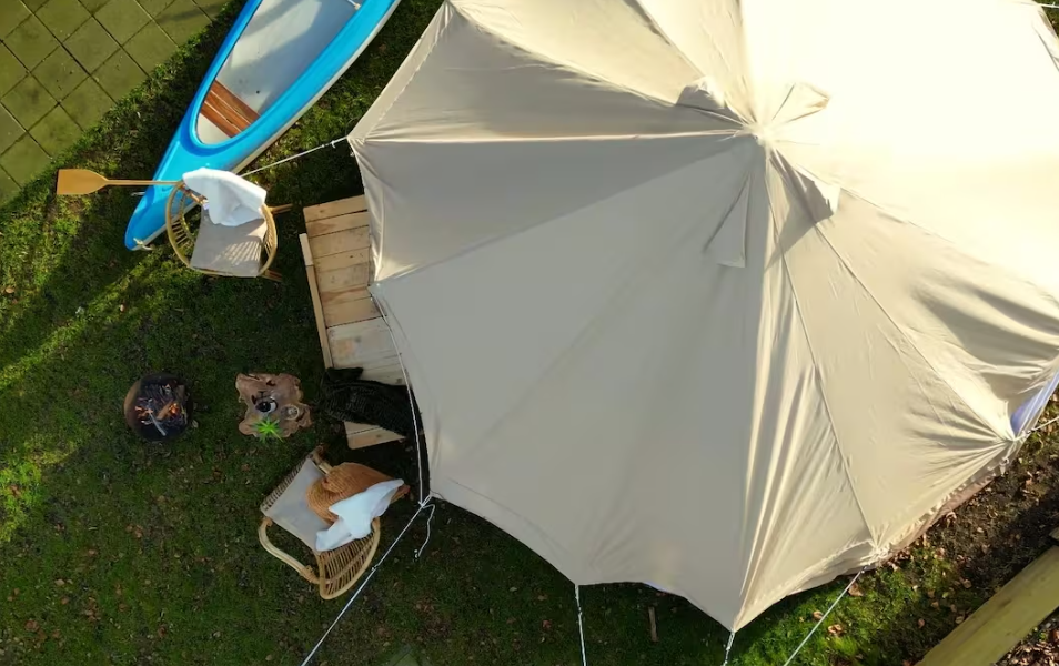 Bovenaanzicht van de beige Sibley ProTech 500 tent van CanvasCamp op de proefslaaplocatie bij Camping Zwolsedijk met een blauwe kano, houten vlonder, stoel en kampvuur ernaast.