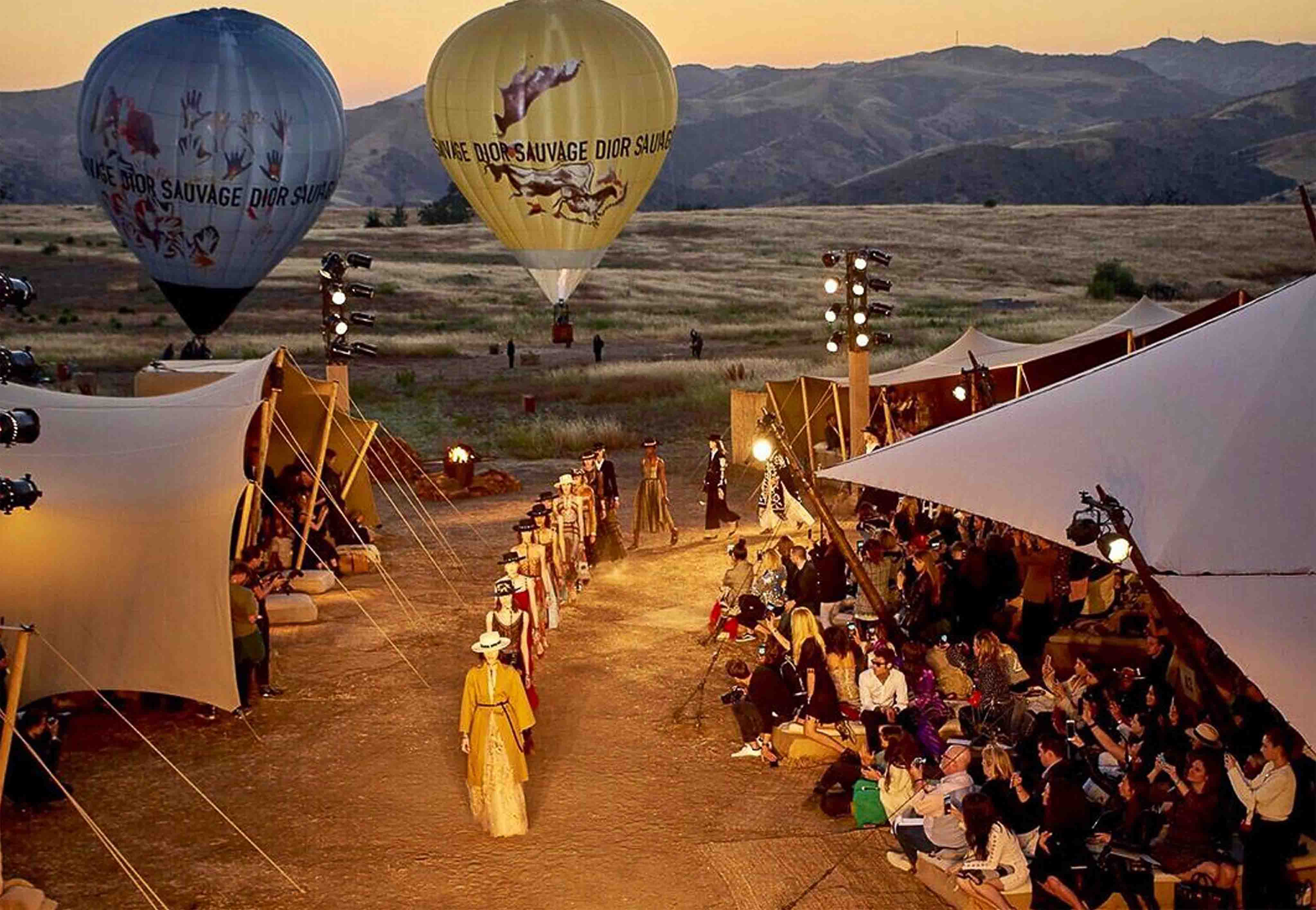 Een openlucht modeshow bij zonsondergang, met modellen die over een zandkleurige catwalk lopen tussen grote stretchtenten vol toeschouwers. Op de achtergrond stijgen twee luchtballonnen op met de tekst "DIOR SAUVAGE" en illustraties. Het evenement vindt plaats in een uitgestrekt, heuvelachtig landschap met gouden grasvelden.
